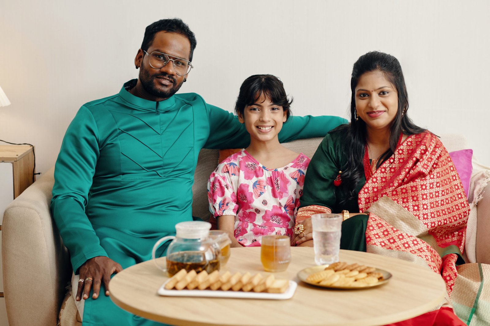 Family of three dressed in traditional attire sitting on a couch smiling together, with snacks and drinks on the table in front of them